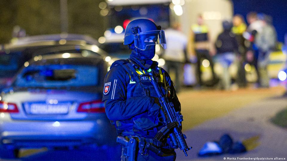 Ein Polizist mit Helm und Maschinenpistole steht neben einem Auto mit zerbrochener Heckscheibe 