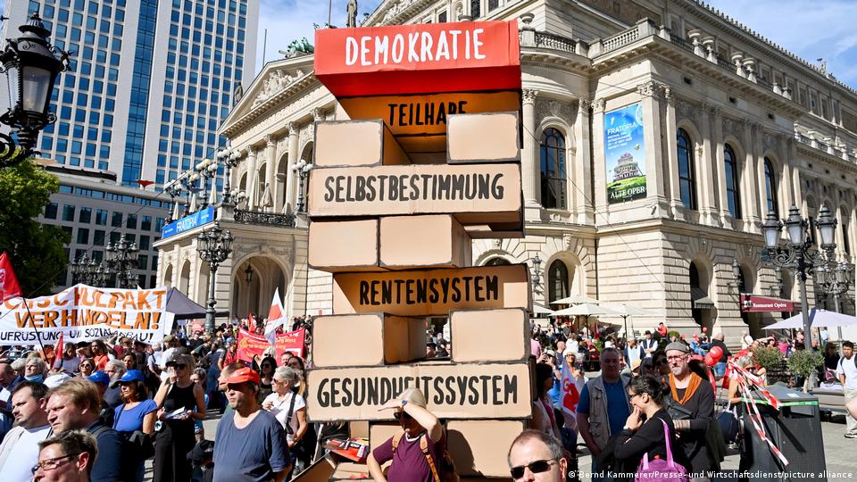 Demonstranten in Frankfurt stellen einen Jenga-Turm mit Blöcken mit der Aufschrift „Teilhabe, Selbstbestimmung, Rentensystem, Gesundheitssystem“ zur Schau, die „Demokratie“ hochhalten.